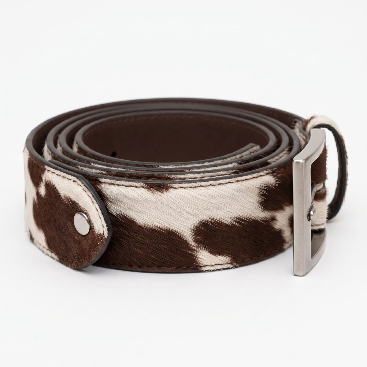 "Brown and white cow-print calf hair leather belt rolled into a spiral coil, showing dark brown leather interior lining and a silver-tone buckle on the right, photographed on a white background."