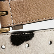 Extreme close-up macro shot comparing two leather belt textures — tan pebbled leather with buckle and stitching on top, and cow-print calf hair leather with black spot and eyelets on bottom."