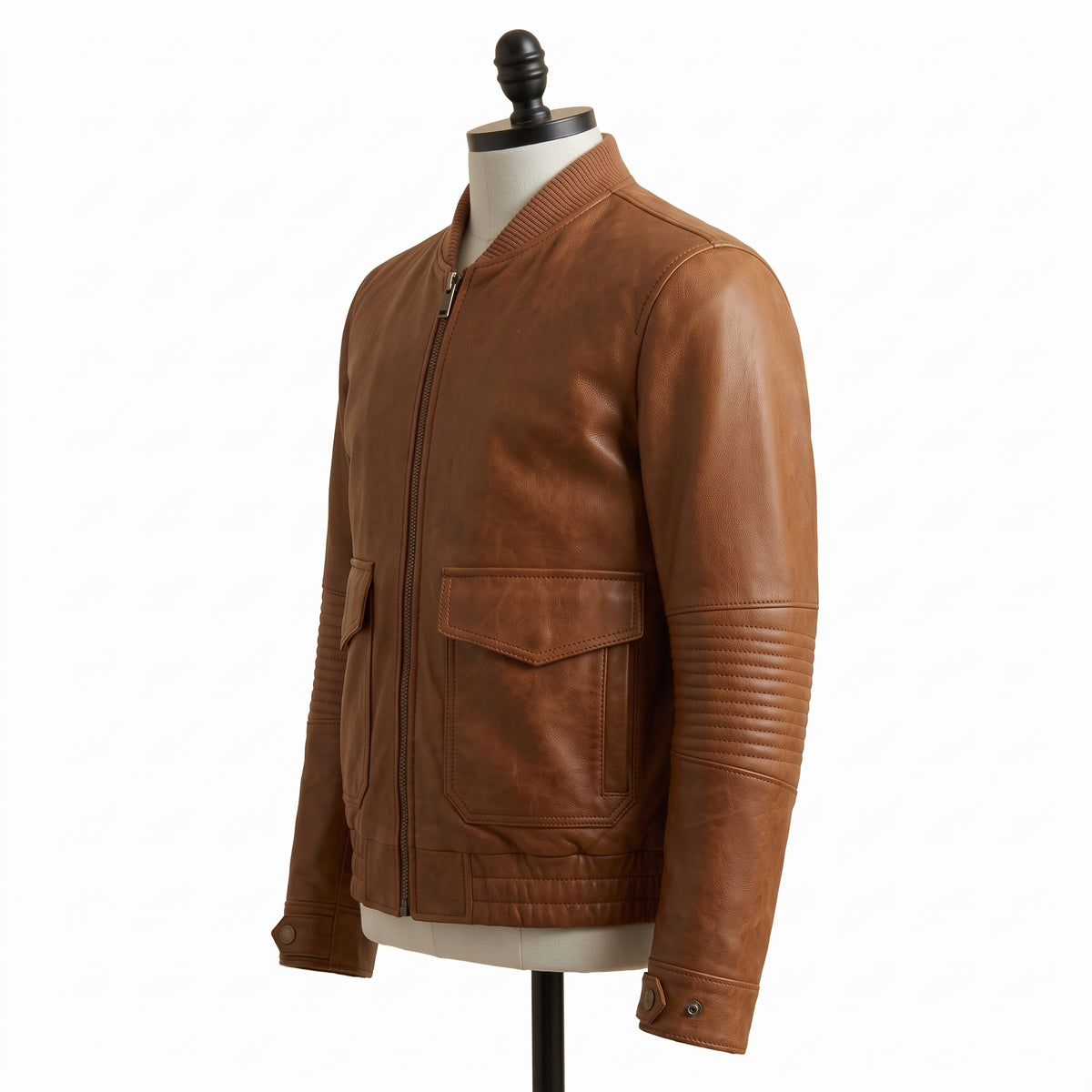 "Angled side view of a tan brown leather bomber jacket with ribbed collar, front zipper, flap pockets, and quilted elbow panels, displayed on a mannequin against a white background."