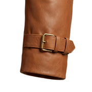 "Close-up of the sleeve cuff on a tan leather trench coat, showing the brass buckle strap and leather grain texture."