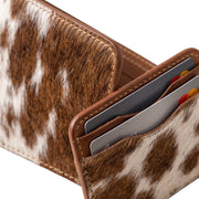 "Close-up top-down view of a cow-print leather bifold wallet standing open beside a matching card holder filled with credit cards, showing stitching and texture detail on a white background." 