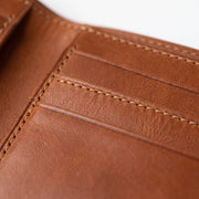"Extreme close-up macro shot of tan brown leather wallet card slots, showing fine pebbled leather grain texture, layered stacked card compartments with visible edges, and contrasting cream/tan stitching running diagonally across the frame, softly blurred background transitioning to white."
