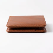 "Low-angle close-up of a tan brown leather bifold wallet lying flat on a white surface, captured from the front edge showing the thin profile, layered leather panels, visible cream stitching along the border, and rich grain texture, photographed with soft studio lighting and a subtle shadow beneath."