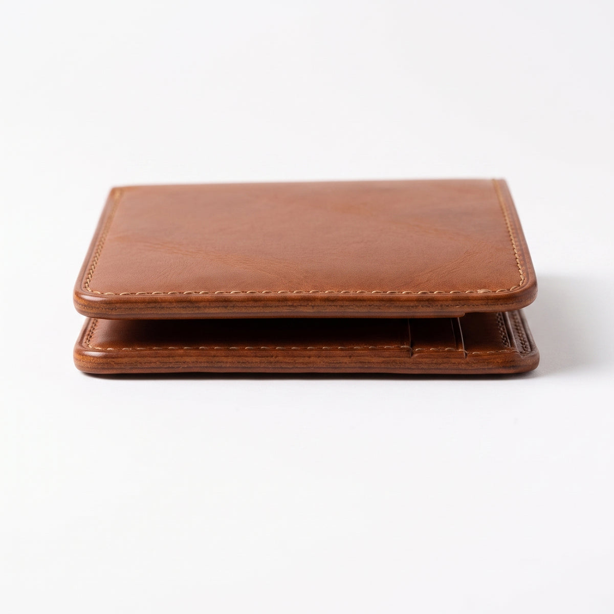 "Low-angle close-up of a tan brown leather bifold wallet lying flat on a white surface, captured from the front edge showing the thin profile, layered leather panels, visible cream stitching along the border, and rich grain texture, photographed with soft studio lighting and a subtle shadow beneath."
