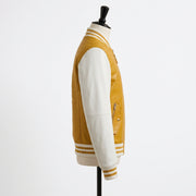 A side-profile view of a varsity-style jacket displayed on a mannequin against a plain white background. The jacket has a textured mustard yellow body with contrasting smooth off-white sleeves and striped ribbed trim.