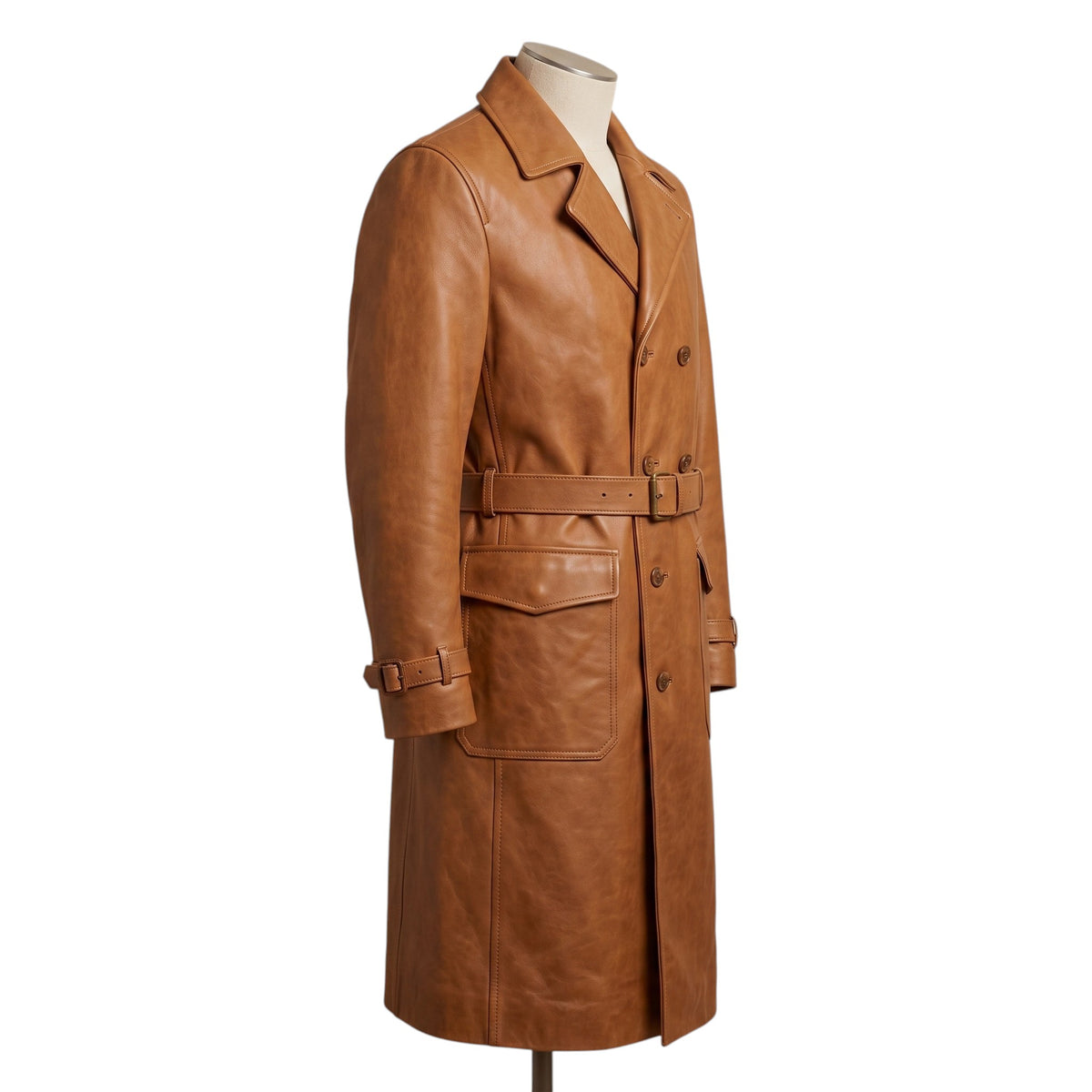 "Another side profile view of the tan leather trench coat on a mannequin, showing the double-breasted button closure, waist belt with brass buckle, and flap pocket against a white background."