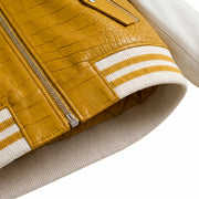 Close-up detail of a varsity-style jacket showing a mustard yellow crocodile-embossed leather panel with a silver zipper, contrasted against cream ribbed knit trim with two white stripes.