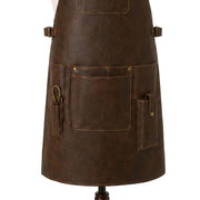 "Close-up of distressed brown leather apron's lower section showing dual waist pockets, a brass scissor loop, side buckle straps, and a tool pocket with snap closure."