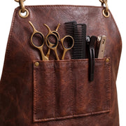 "Close-up of brown leather apron pocket holding gold scissors, comb, clip, and clipper tool."