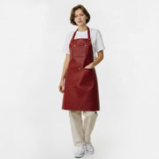 A young woman with short brown hair wearing a red leather bib apron over a white t-shirt and cream-colored wide-leg trousers, paired with white sneakers. The apron features a square chest pocket and a wide front pocket with brass snap buttons, styled against a plain white background."