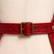 "Extreme close-up of a red leather apron's back waist strap, showing a rectangular brass buckle threaded through the belt with multiple adjustment holes, secured to the apron's side panels with brass rivets, against a cream-colored dress form mannequin background.