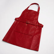 "Flat lay of a red leather bib apron laid diagonally on a white background. Shows the full apron shape including the neck strap, square chest pocket, side waist buckle, and wide bottom pocket with a center divider, all detailed with brass rivets and snap buttons on textured red leather."