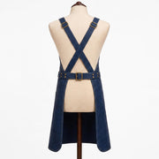"Back view of navy blue leather apron on a fabric mannequin, showing crossback straps with brass buckles, an adjustable waist belt with a center buckle, split hem design, and contrast stitching throughout."