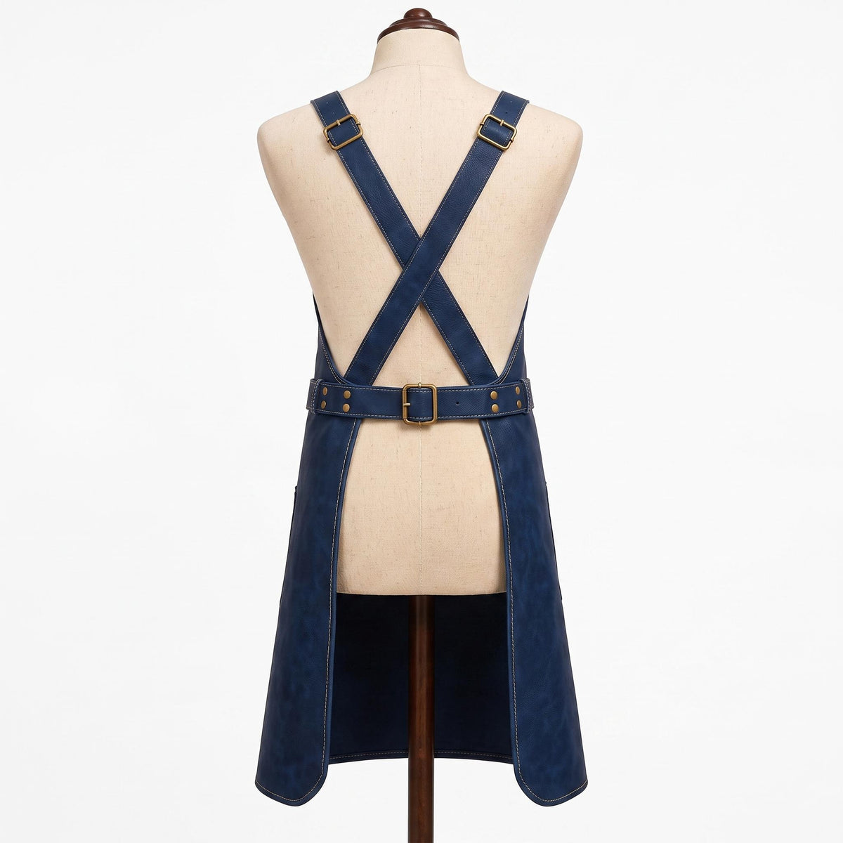 "Back view of navy blue leather apron on a fabric mannequin, showing crossback straps with brass buckles, an adjustable waist belt with a center buckle, split hem design, and contrast stitching throughout."