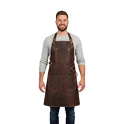 "Smiling man wearing a grey sweater and dark brown leather apron with adjustable neck straps, chest pocket, and multi-compartment lower pocket, standing against a plain white background."