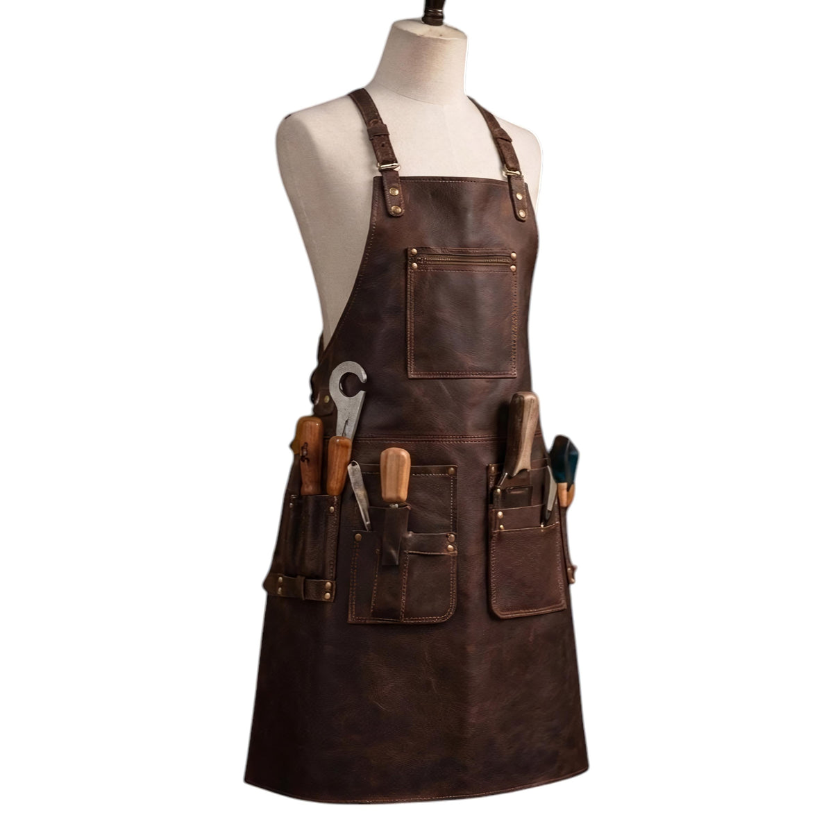 "Front angled view of a dark brown leather work apron displayed on a fabric mannequin, featuring adjustable neck straps, a zippered chest pocket, and multiple front tool pockets holding various tools including a wrench, chisels, wooden-handled hand tools, and a screwdriver, photographed against a plain white background."