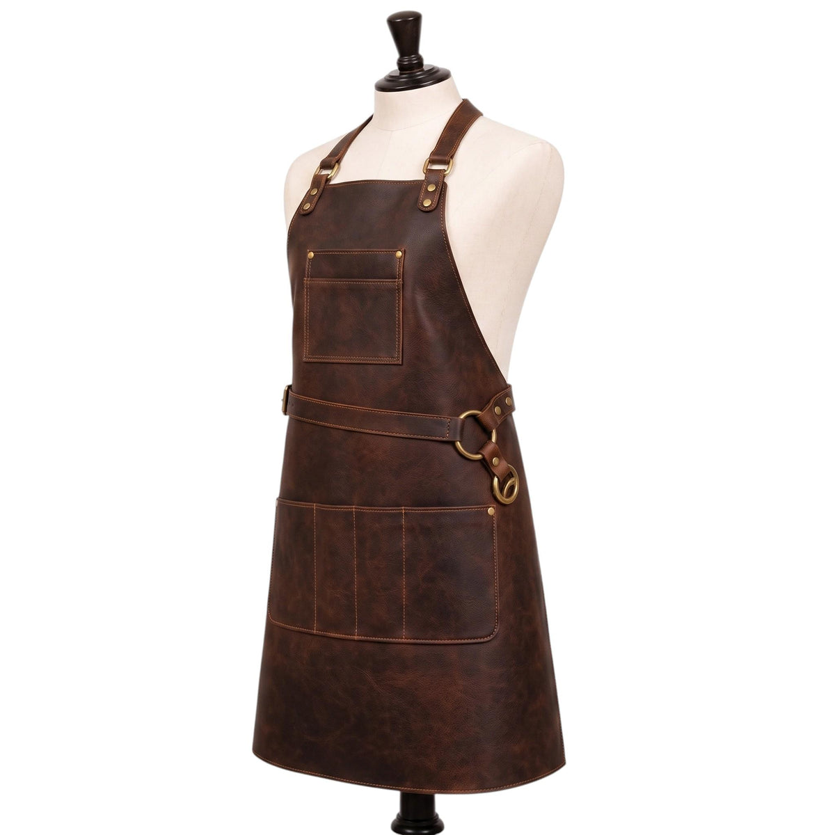 "Angled front view of a dark brown leather apron on a cream mannequin torso, showing an adjustable neck strap with brass buckles and D-rings, a diagonal waist strap with brass ring hardware, a chest pocket, and a wide multi-compartment lower pocket, against a plain white background."