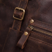 Close-up of a dark brown distressed leather bag showing a brass buckle strap, a zippered pocket, and brass rivets.