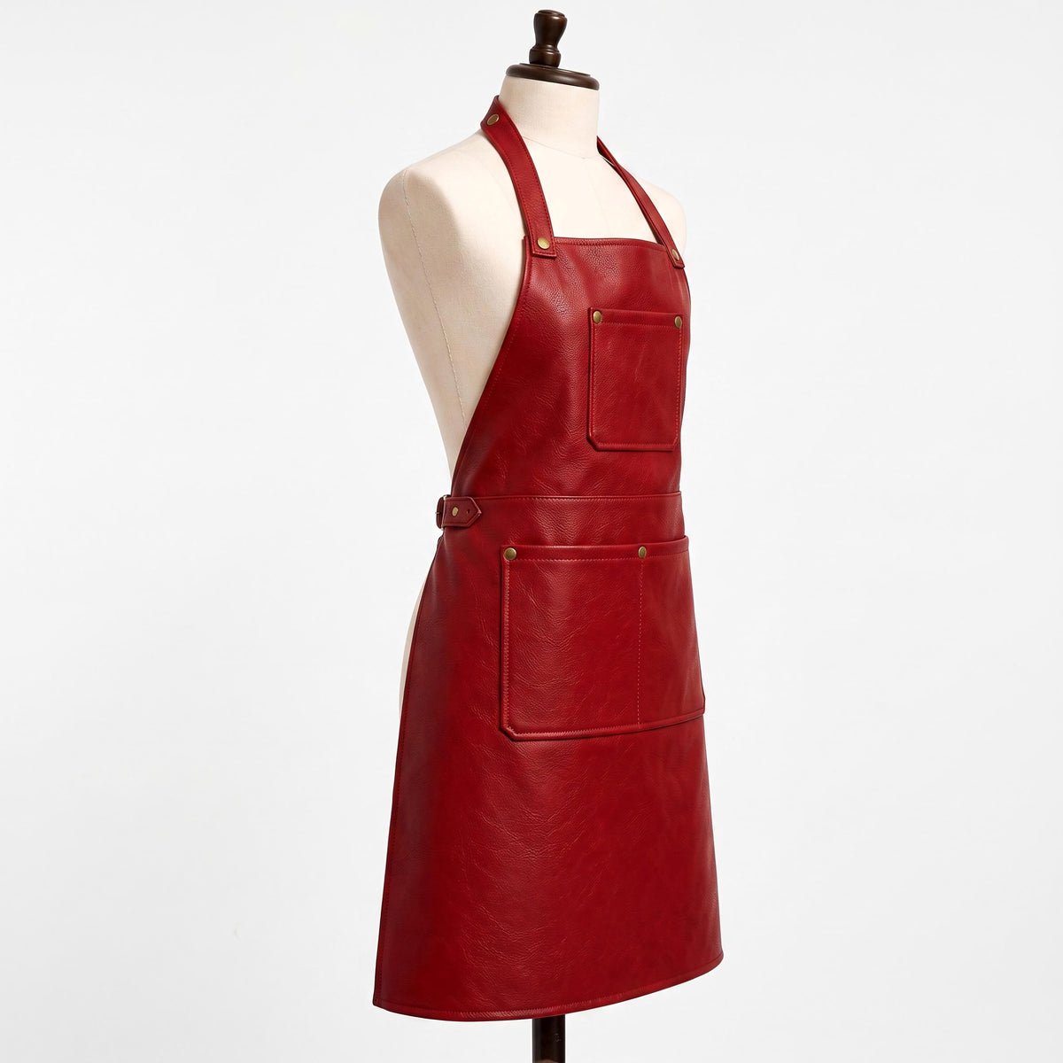 "Side profile view of a red leather bib apron on a cream-colored dress form mannequin against a white background. Shows the halter neck strap secured with a brass rivet, a square chest pocket, an adjustable side buckle at the waist, and a wide lower pocket with brass snap buttons