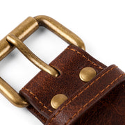 Close-up of a brown leather belt with an antique brass buckle and matching brass rivets, shown on a white background.