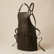 Three-quarter product view of dark brown leather workshop apron with adjustable crossback harness, front pockets and side tool holders.