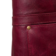 "Close-up detail of burgundy leather texture showing a waist strap and a brass snap-button pocket edge."