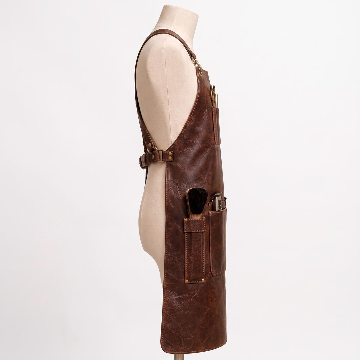 "Side view of brown leather barber apron on mannequin, showing pockets with scissors, comb, brush, and clippers."