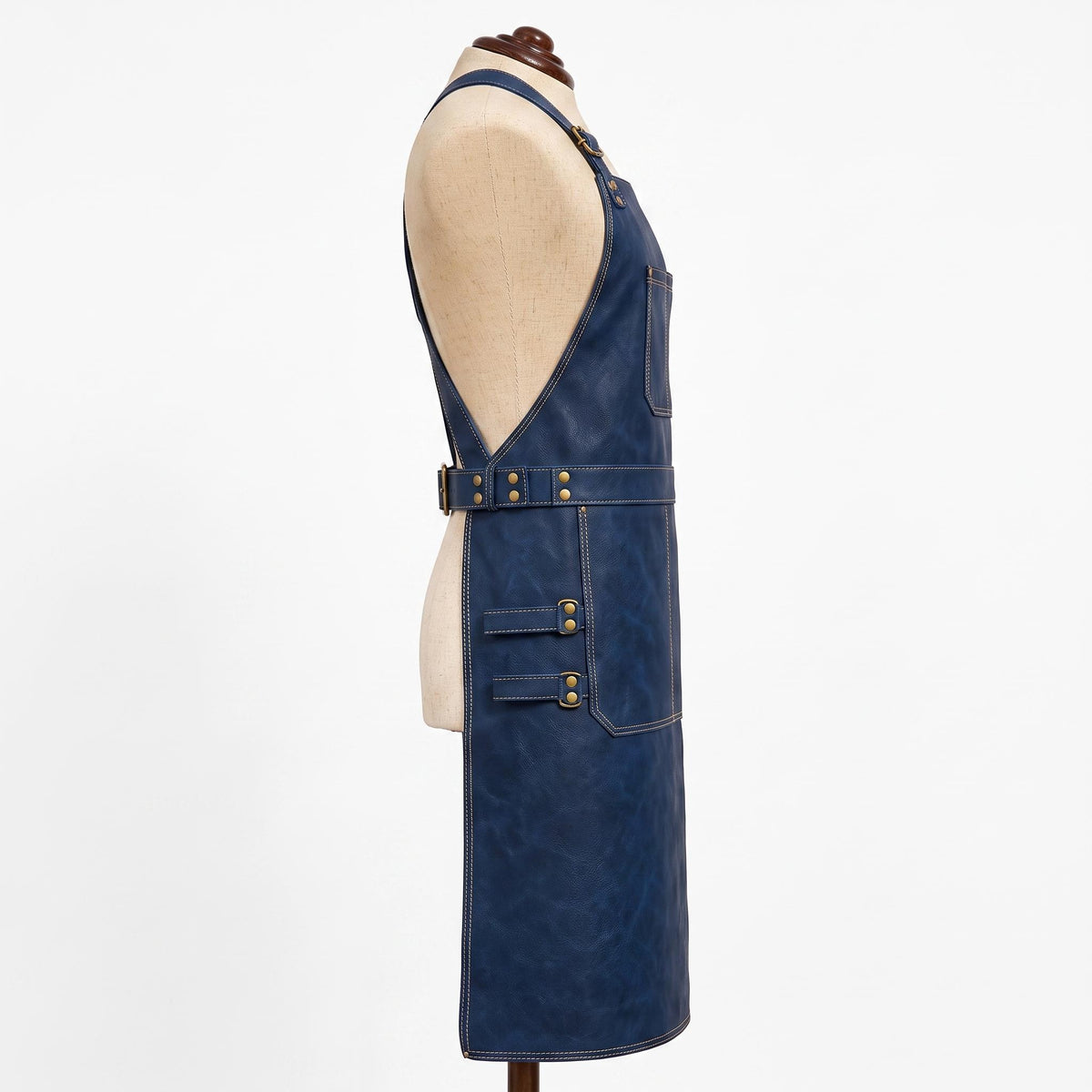 "Profile view of navy blue leather apron on a mannequin, showing the deep armhole cut, side chest pocket, adjustable waist belt with brass buckle, and a lower pocket with two side tool loops."