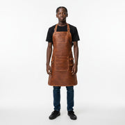 "Young model wearing black t-shirt and jeans, dressed in brown distressed leather work apron with adjustable neck strap, chest pocket, and front pocket with D-ring, standing against a plain light background."