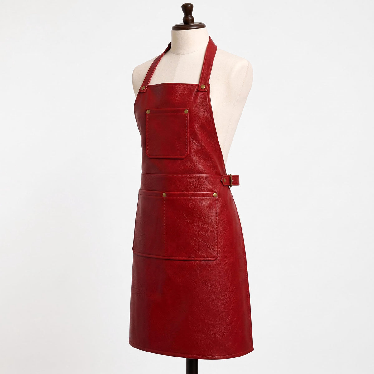 "Angled side view of a red leather bib apron on a cream-colored dress form mannequin against a white background. Shows the halter neck strap with brass rivets, a square chest pocket, a brass side buckle for waist adjustment, and a wide front pocket with brass snap buttons near the hem."