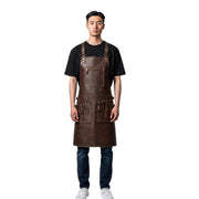 A man wearing a dark brown leather work apron over a black t-shirt, blue jeans, and white sneakers, standing against a solid white background.