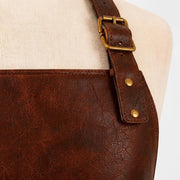 "Close-up detail of a brown leather apron's shoulder strap, showing the antique brass buckle, adjustment holes, and riveted stitching where the strap attaches to the apron body."