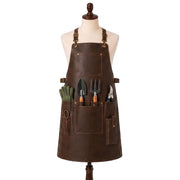 "Distressed brown leather gardening apron on a mannequin with tool pockets holding a trowel, hand fork, pruning shears, and green gardening gloves."