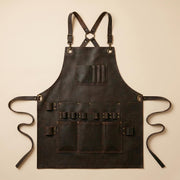 Flat lay of brown leather woodworking apron showing full apron shape, cross-back harness, long waist straps, tool loops and utility pockets.