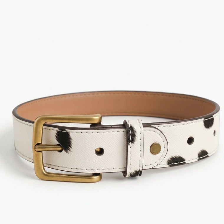 "Cow-print (white with black spots) leather belt formed into a circular loop, showing an antique brass buckle, tan interior lining, and white stitching, photographed on a white background."