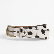 "White leather belt with black spotted (dalmatian/cow-print) pattern, featuring an antique brass buckle and white stitching detail, coiled and photographed on a plain white background."