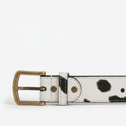 Front-on close-up of a cow-print leather belt buckle in antique brass finish, showing the belt loop, rivets, and stitching detail against a white background."