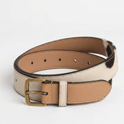 "Reversible leather belt coiled to show both sides — tan/beige smooth leather on one side and cow-print (white with black spots) on the other — with an antique brass buckle, photographed on a white background."