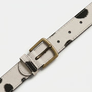 "Close-up diagonal view of a white leather belt with black spotted (cow-print) pattern, showing an antique brass buckle, belt loop, and stitching detail in sharp focus against a white background."