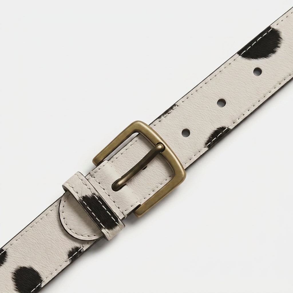 "Close-up diagonal view of a white leather belt with black spotted (cow-print) pattern, showing an antique brass buckle, belt loop, and stitching detail in sharp focus against a white background."