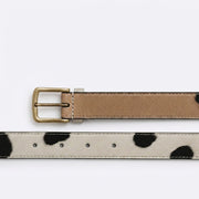 "Two reversible leather belts laid parallel on a white background — top belt showing tan smooth leather side with brass buckle, bottom belt showing cow-print (white with black spots) side with stitching detail."