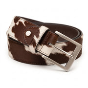 : "Close-up angled view of a brown and white cow-print calf hair leather belt coiled to display a brushed silver-tone buckle, rivet, and stitching detail against a white background."
