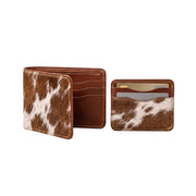 "Half-open cow-print (brown and white) leather bifold wallet showing card slots and 'Handcrafted' embossing on the left, and a matching card holder with credit cards inside on the right, both photographed on a white background."
