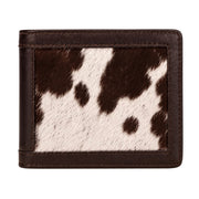 "Front-facing view of a closed cow-print (brown and white) leather bifold wallet with dark brown leather trim and stitching, photographed on a white background."